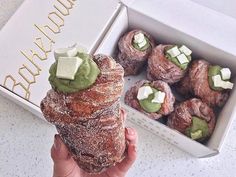 a person holding up a pastry in front of a box of pastries with green toppings