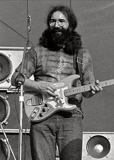 black and white photo of man playing guitar on stage with speaker equipment in the background