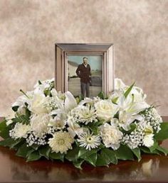 an arrangement of white flowers and greenery sits on a table next to a framed photograph