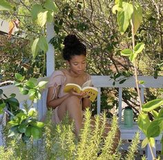 a woman sitting on a bench reading a book in the shade with trees and bushes around her