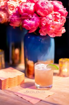 pink flowers are in a blue vase next to a wooden table with coasters and napkins