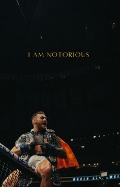 a man sitting on top of a wrestling ring holding a red and yellow flag with the words i am notrious above him