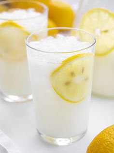 two glasses filled with lemonade and ice on top of a white table next to sliced lemons