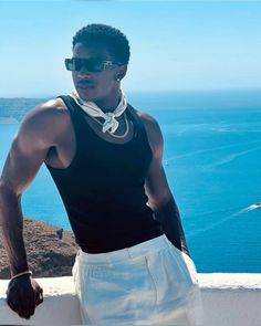 a man wearing sunglasses and a black tank top stands on a ledge near the ocean