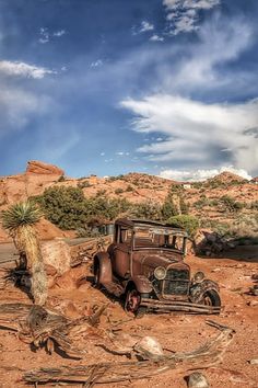 an old car is parked in the dirt near some rocks and cactus trees, under a blue sky with wispy clouds