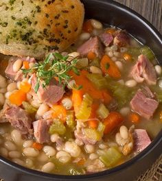 a bowl of soup with ham, beans and bread on a wooden table next to a spoon