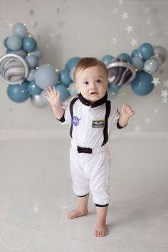 a baby in an astronaut suit standing on the floor with his hands up and looking at the camera