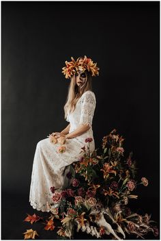 a woman with flowers in her hair sitting on a pile of dead plants and leaves