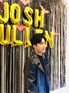 a young man standing in front of a wall with gold foil balloons that spell out the word hush