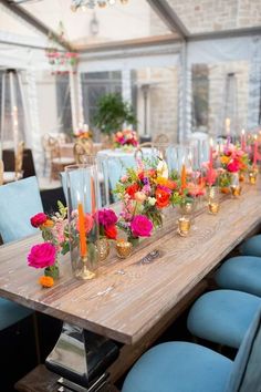 a long wooden table topped with lots of colorful flowers and candles next to blue chairs