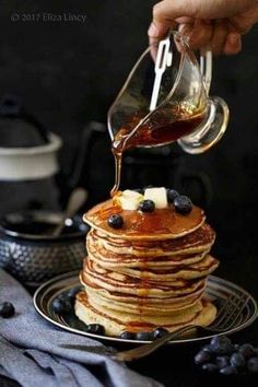 a stack of pancakes with syrup being poured onto them and blueberries on the side