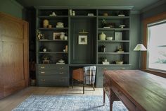 a room with green bookcases and wooden desk in front of a large window