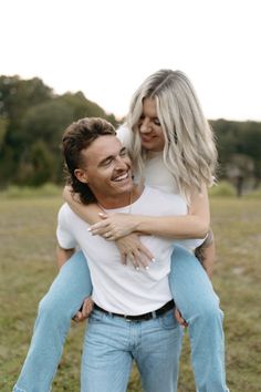 a man holding a woman on his back in the middle of an open field with trees in the background