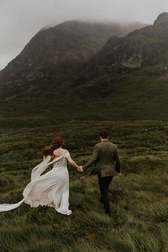 a bride and groom holding hands while walking through the grass with mountains in the background