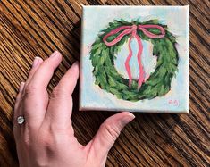 a hand is holding up a small canvas with a wreath painted on it and pink ribbon