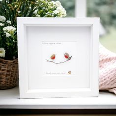 two little birds sitting on a branch in a white frame next to a basket with flowers