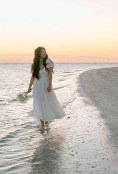 a woman in a white dress is walking on the beach at sunset with her hair blowing back