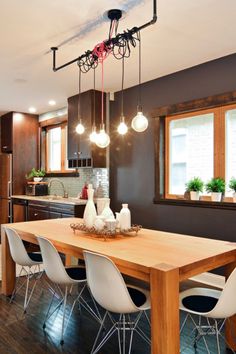 a dining room table with white chairs and lights hanging from the ceiling above it, in front of an open kitchen area