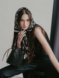 a woman sitting on a chair holding a black purse and looking at the camera while wearing a scarf over her head