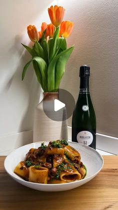 a white plate topped with pasta next to a bottle of wine and vase filled with orange flowers