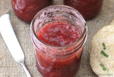 three jars filled with strawberry jam next to a cookie on a cloth and two spoons