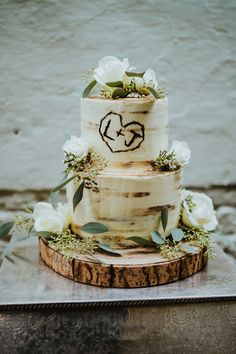 a three tiered wedding cake with white flowers and greenery on top, sitting on a wooden slab