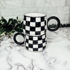 two black and white checkered coffee mugs sitting on a marble counter next to green plants