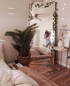 a woman standing in front of a large mirror next to a bed and plant on the floor