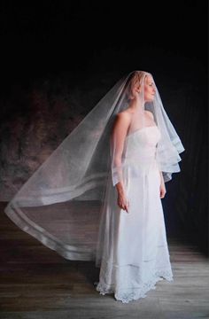 a woman in a white dress and veil standing on a wooden floor with dark background