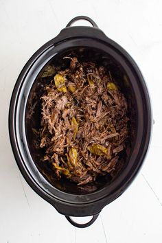 shredded meat in a slow cooker on a white counter top with peppers and olives