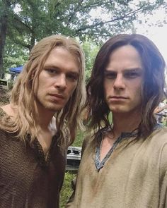 two men standing next to each other in front of trees and grass, one with long hair