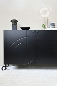 a black sideboard with wheels and a vase on the top, in front of a white wall