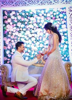 a man kneeling down next to a woman in front of a flower covered wall with flowers on it