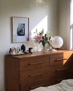 a dresser with flowers on it in front of a window and an art piece hanging above