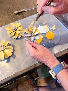 a person painting flowers on a table with yellow and white paint, while another hand holds a brush