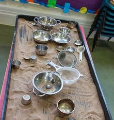 there are many silver bowls and spoons on the sand covered picnic table with chairs in the background