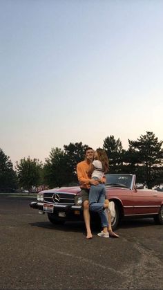 a man and woman sitting on the hood of a vintage car in a parking lot