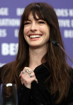 a smiling woman sitting at a table with a glass in front of her and a microphone behind her