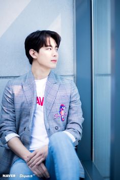 a young man sitting on top of a bench next to a wall wearing a blazer and jeans