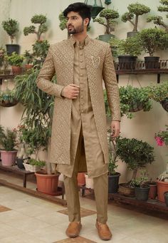 a man standing in front of potted plants wearing a tan sherwanit