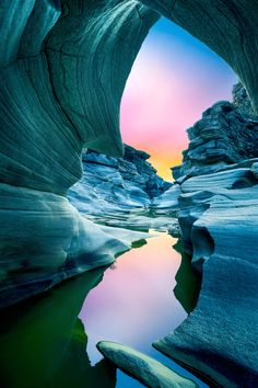 the view from inside an ice cave looking down at water and rocks, with a colorful sky in the background