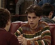 a young man sitting at a table talking to another person in front of him with other people behind him