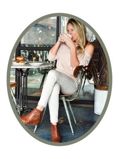 a woman sitting at a table drinking from a cup with her legs crossed and wearing white pants