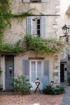 an old stone building with blue shutters and flowers on the outside wall, next to a brick walkway