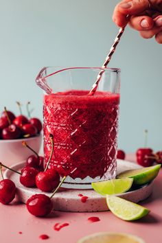 someone holding a straw over a glass filled with red liquid and cherries on the side