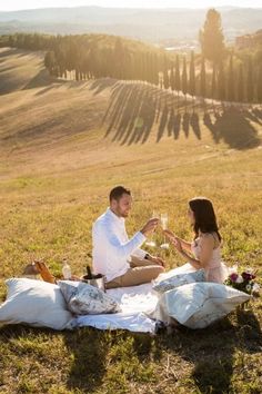 a man and woman sitting on top of a grass covered field holding glasses of wine