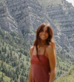 a woman standing in front of a mountain with trees on the side and mountains behind her