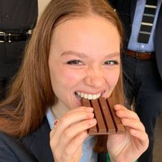 a woman holding up a chocolate bar to her face while wearing a suit and green beaded necklace