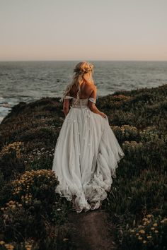 a woman in a white dress is walking along the grass by the ocean with her back to the camera