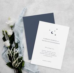 the wedding card is next to some flowers on top of a white table cloth and blue paper